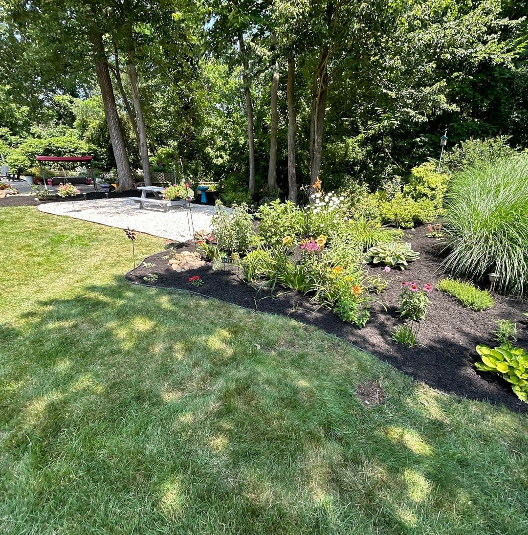 Lush garden landscape with vibrant flowers, trees, and a gravel patio area.