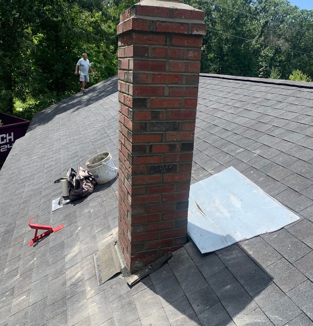 Chimney on a roof with tools and a worker in the background surrounded by trees.
