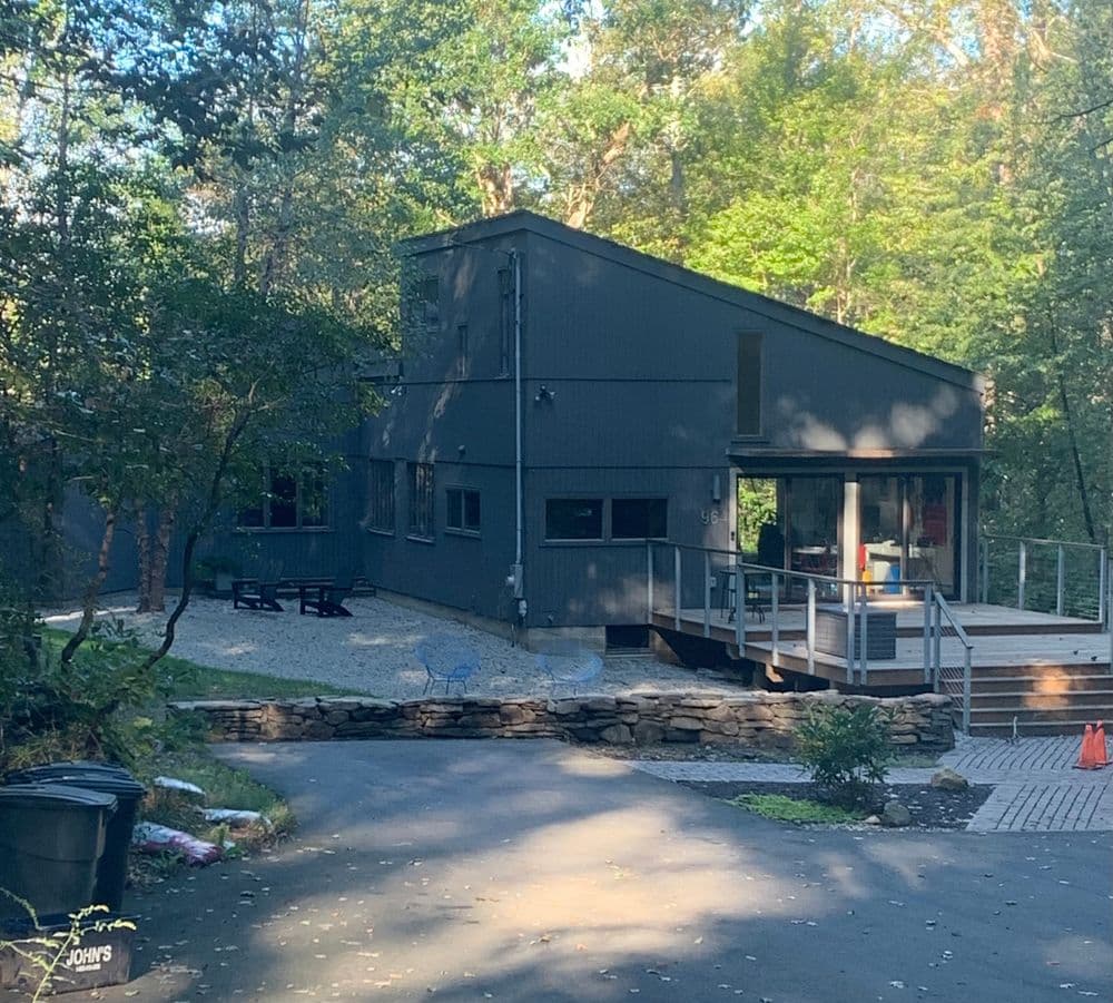 Modern black house with wooden deck surrounded by trees and gravel driveway.