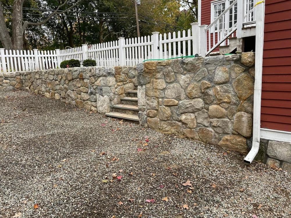 Stone retaining wall with stairs in gravel yard next to a red house and white picket fence.