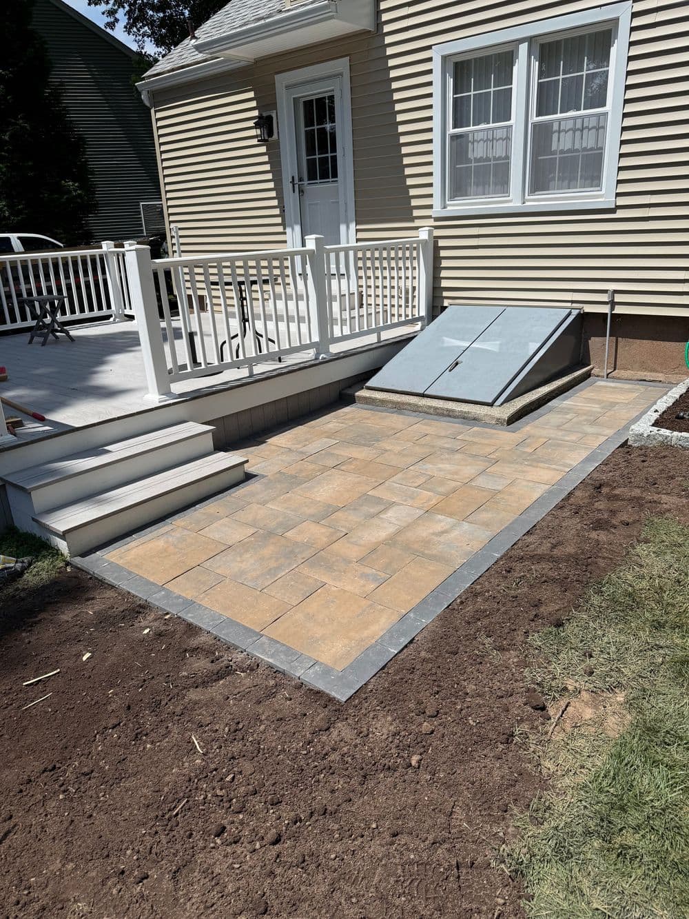 Patio installation with stone pavers and a deck area next to a beige house exterior.