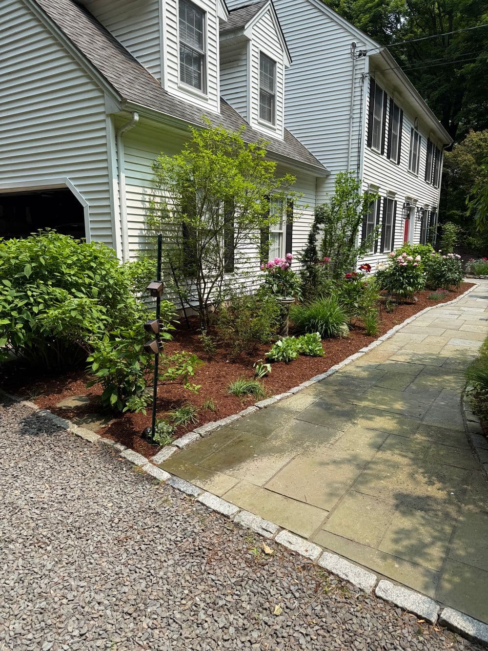 Lush garden with shrubs and flowers alongside a stone pathway by a white house.