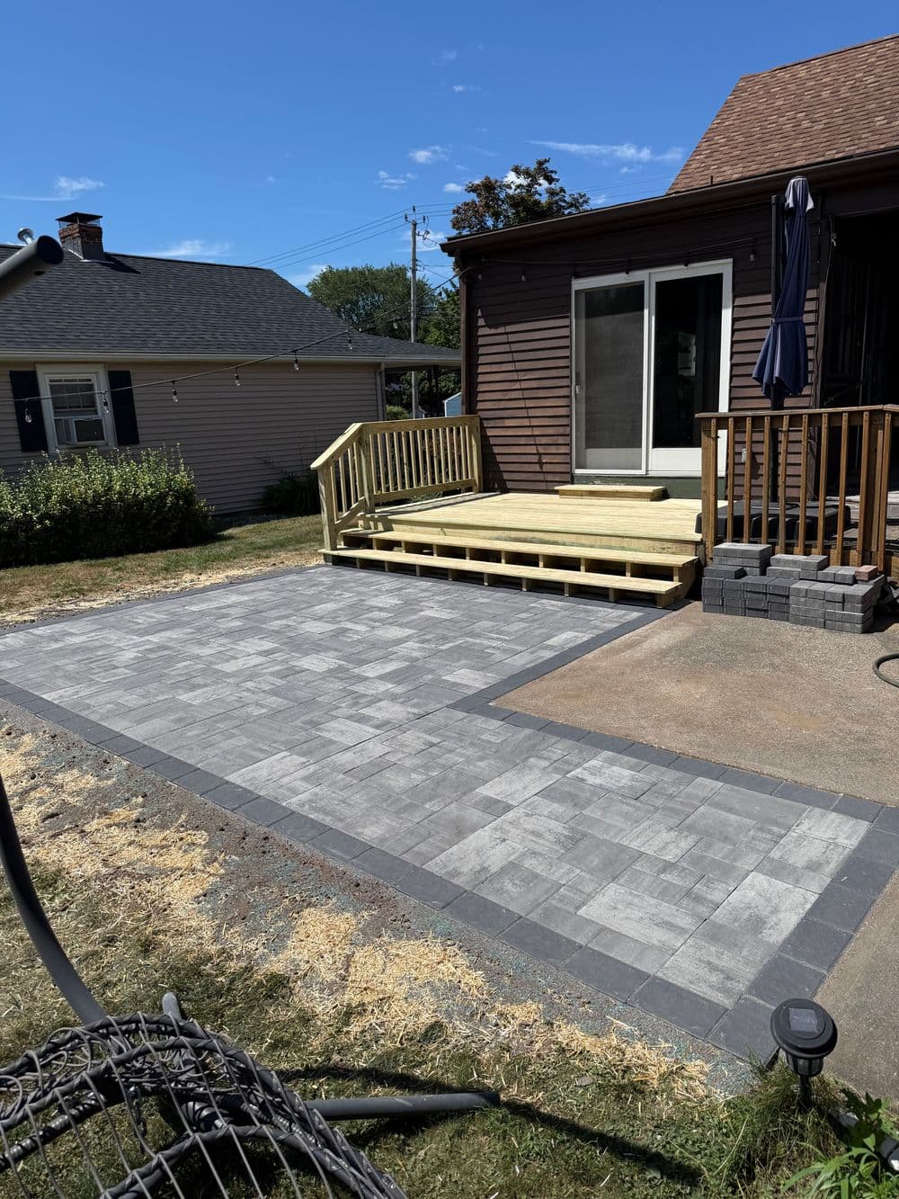 Newly installed patio with gray pavers, wooden deck, and clear blue sky in the background.